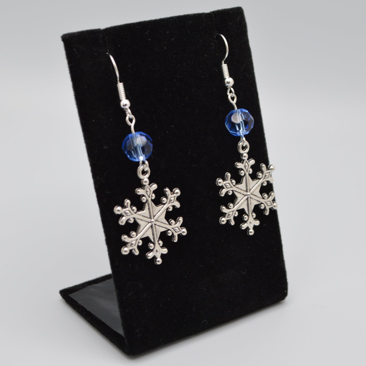 Silver snowflake earrings with blue beads on a black display stand.