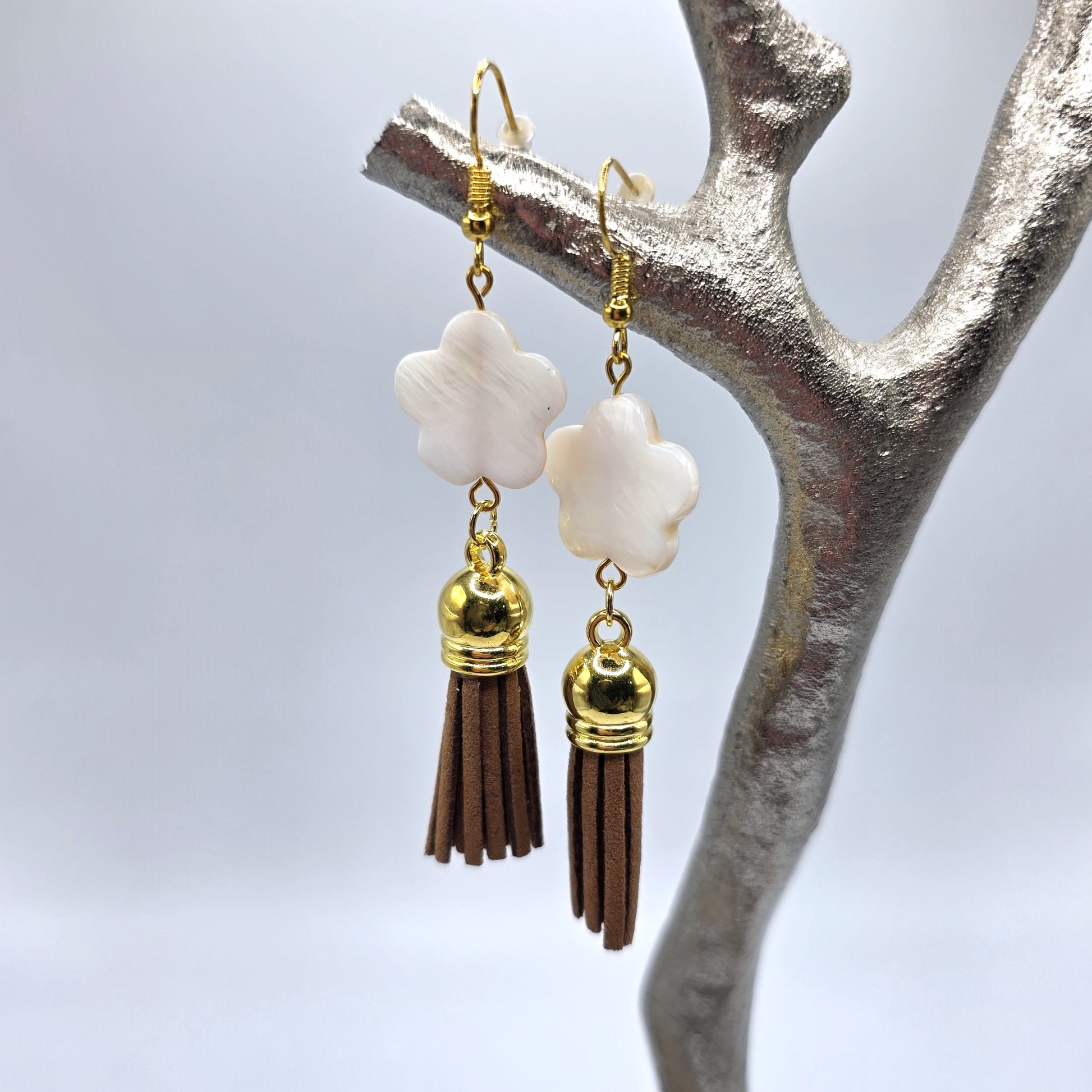 Pair of earrings with white flower-like beads and brown tassels hanging on a branch.
