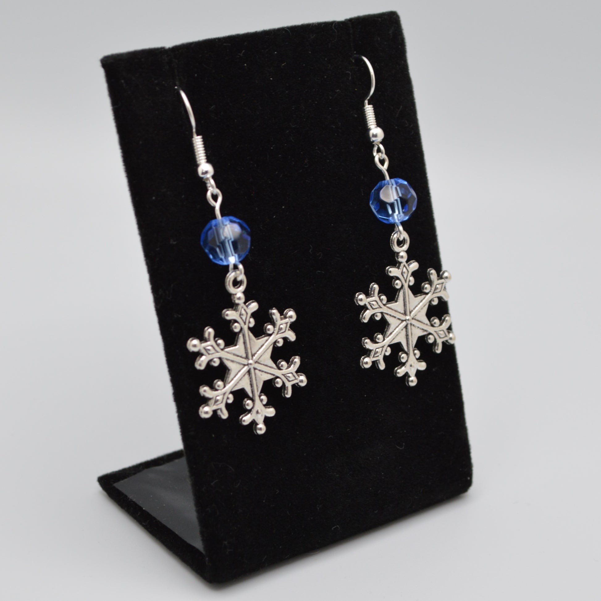 Silver snowflake earrings with blue beads on a black display stand.
